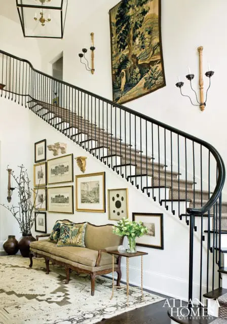 Foyer with a curved staircase featuring a black railing and patterned runner, a tan upholstered bench, a large tapestry, and a gallery wall of framed art.
