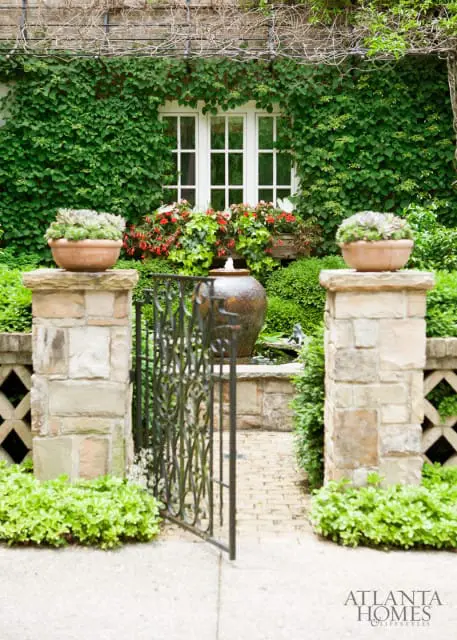 Stone pillars flank an open wrought iron gate leading to a brick path. A large ceramic fountain sits in the background, surrounded by lush greenery and a window covered in ivy.