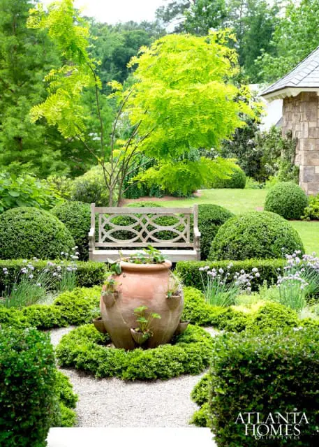 Formal garden with manicured boxwood hedges, a terracotta strawberry pot, a wooden bench, and a stone building in the background.