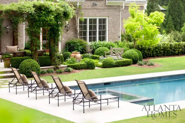 Backyard with a swimming pool, lounge chairs, manicured hedges, a wooden bench, and a brick house with a vine-covered pergola.