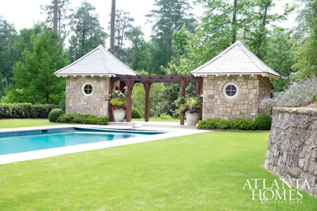 Stone pool house with a pergola connecting two structures, circular windows, and potted flowers, next to a swimming pool and manicured lawn.