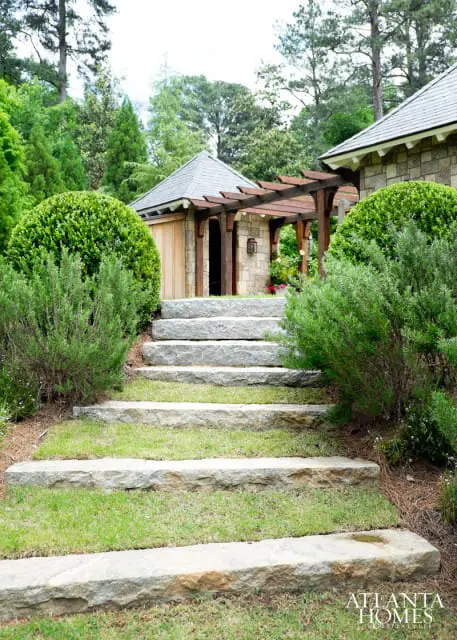 Stone steps leading up to a stone building with a wooden pergola and a grey shingled roof, surrounded by lush green bushes and trees.
