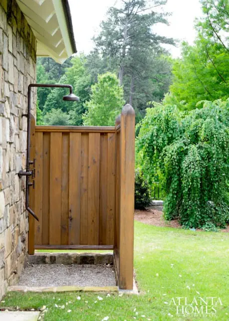 Outdoor shower with rustic metal fixtures attached to a stone wall, adjacent to a wooden privacy screen and lush green landscaping.