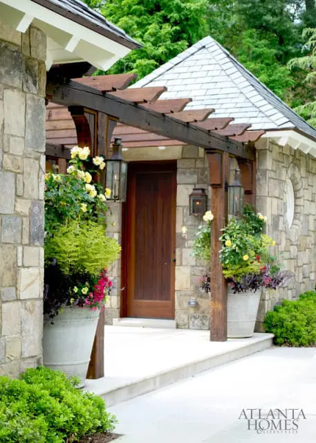 Stone house entrance with wooden pergola, large planters filled with yellow roses and greenery, and a dark wood door.