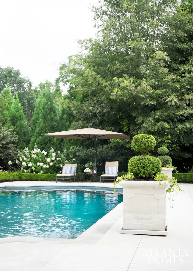 Turquoise swimming pool with white tiled edge, flanked by lounge chairs under a large umbrella, surrounded by lush green trees and manicured hedges.