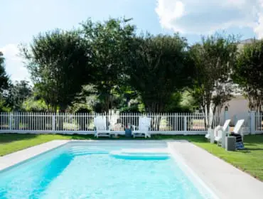 Rectangular swimming pool with white lounge chairs and a white picket fence surrounding a green lawn with trees.