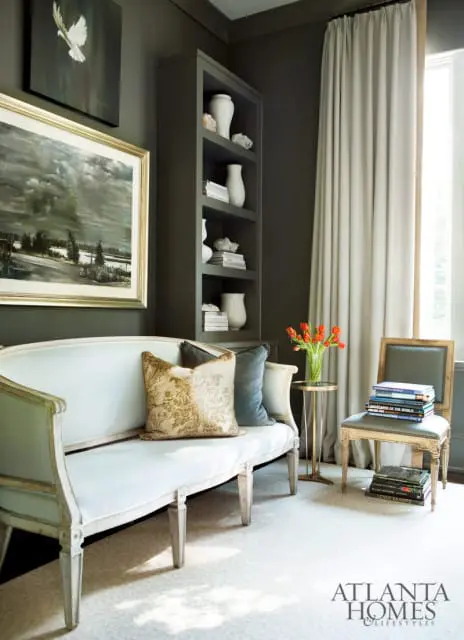 Living room with a light blue settee, dark gray walls, built-in shelves with vases, and a chair stacked with books.