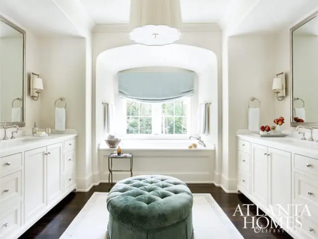 Creamy white bathroom with a freestanding tub under an arched window, double vanities with marble countertops, and a teal tufted ottoman.