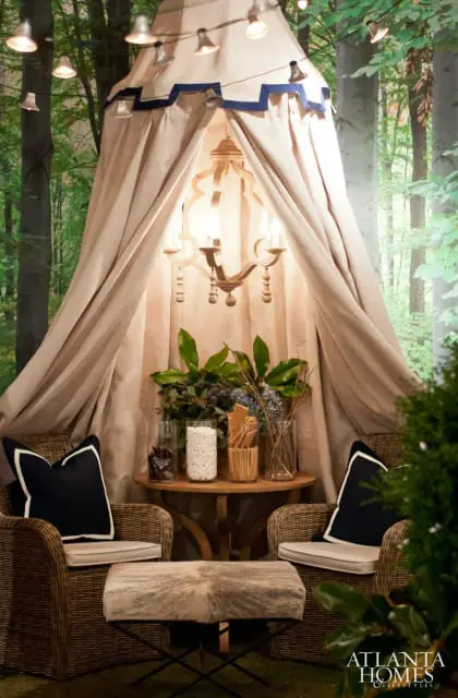 Tented seating area with two wicker chairs, a cowhide ottoman, and a wooden chandelier, set against a forest mural and string lights.