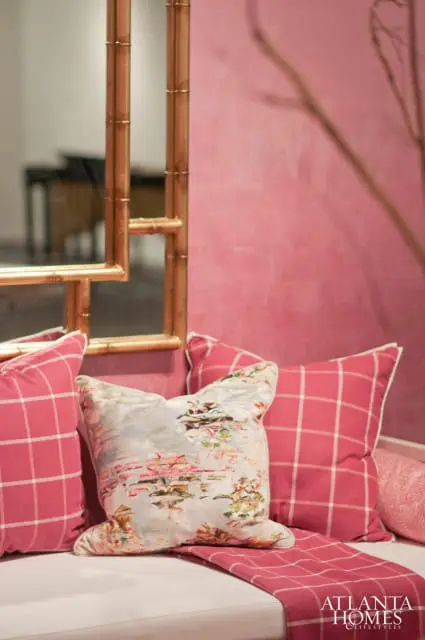 Pink wall with a bamboo-style mirror frame, a sofa with pink plaid pillows, a floral patterned pillow, and a pink plaid throw.