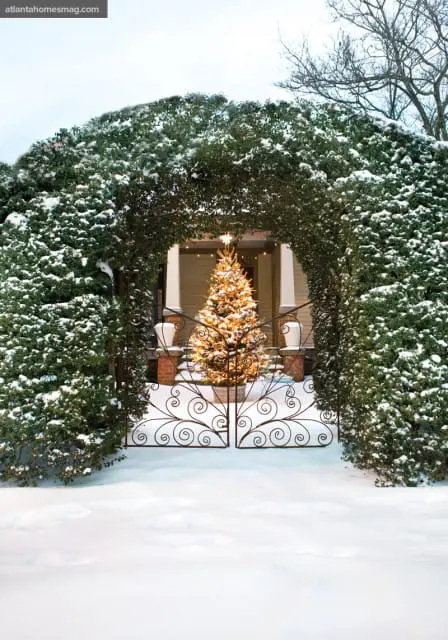 Snow-covered evergreen bushes form an archway over a decorative wrought iron gate, framing a lit Christmas tree in front of a house