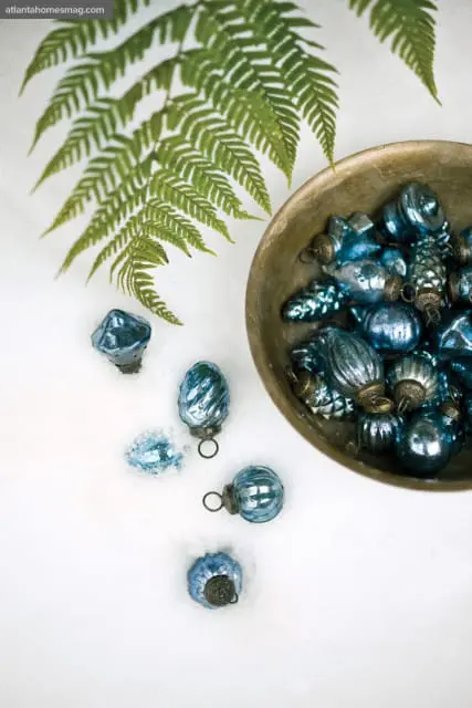 Assortment of vintage blue glass Christmas ornaments in a rustic bowl and scattered in snow, with a green fern frond overhead.