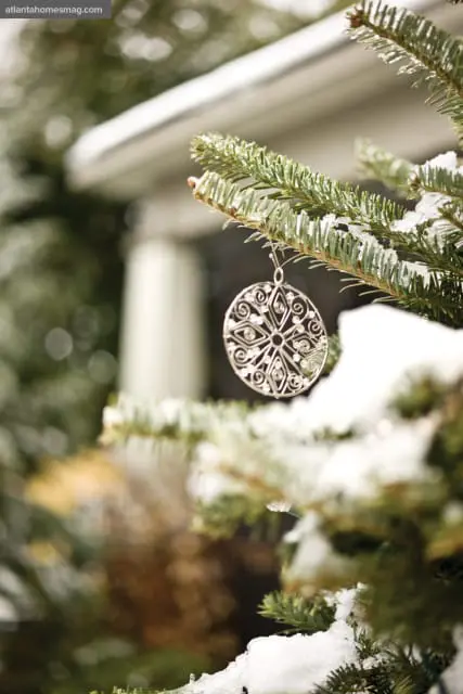 Close-up of a silver snowflake ornament hanging from a snow-dusted pine branch with a blurred white porch column in the background.