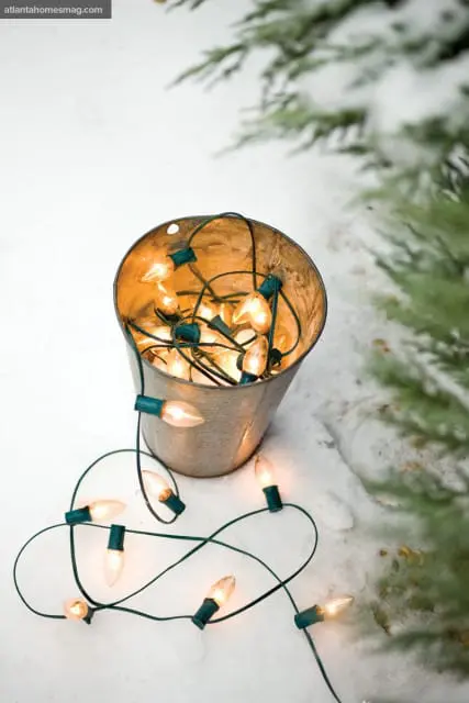 String of clear Christmas lights with green cords spilling out of a galvanized metal bucket in the snow, with a pine tree branch in the background.