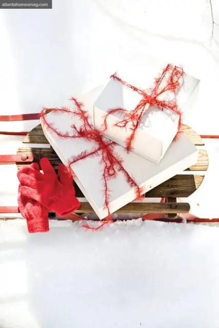 Two white gift boxes tied with red fuzzy ribbon sit on a wooden sled with red runners, next to a pair of red knitted gloves, all on a bed of snow.