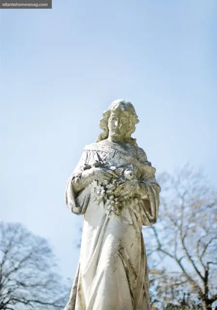 Weathered stone statue of a woman holding a bouquet of flowers, set against a clear blue sky and bare trees.