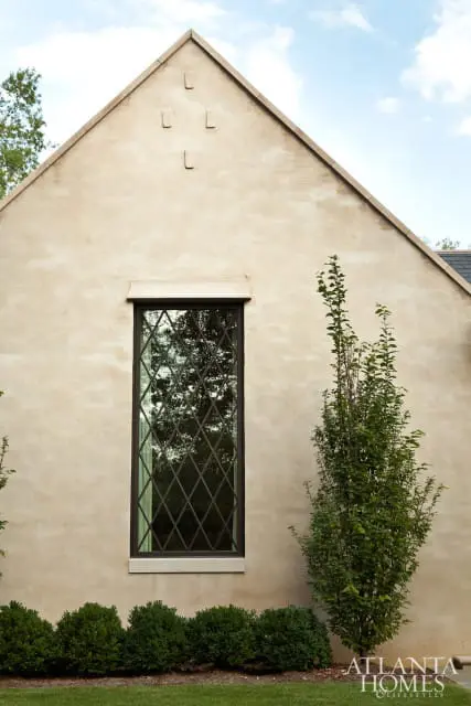 Stucco exterior wall with a tall, narrow diamond-paned window and a small tree.