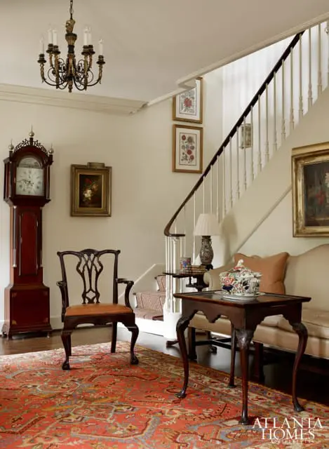 Foyer with a grandfather clock, antique chair, ornate rug, and staircase with floral artwork. A sofa and side table with a decorative tureen are visible.