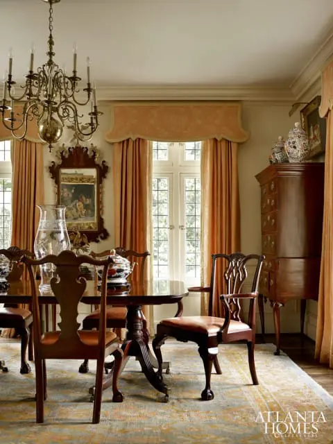 Traditional dining room with dark wood pedestal table and chairs with leather seats, ornate brass chandelier, and peach floral curtains.