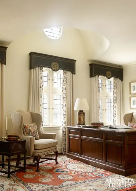 Traditional home office with a large wooden desk, wingback chair, and ornate rug. Cream walls and arched ceiling with a circular window.