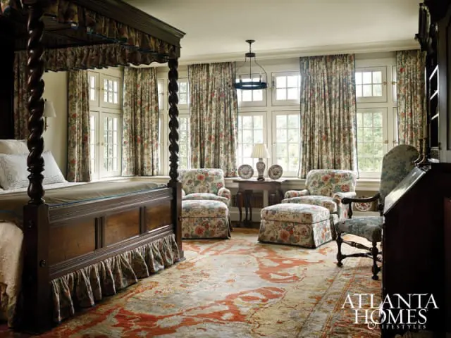 Bedroom with a dark wood four-poster bed, floral upholstered chairs and ottoman, patterned curtains, and a large oriental rug.