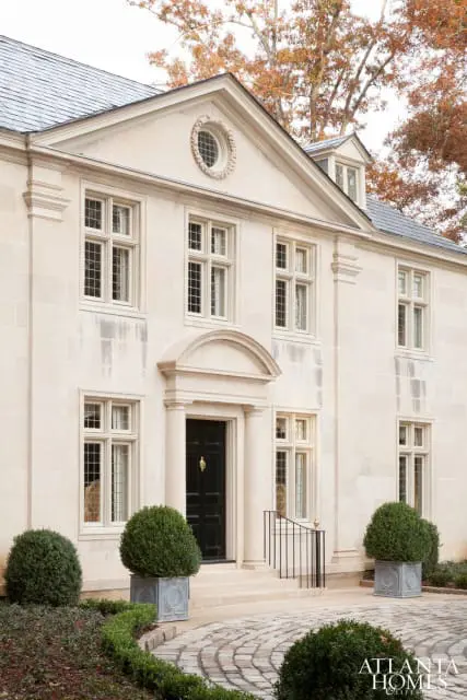 Stone facade of a large home with a black front door, arched entryway, multiple windows, and manicured bushes in square planters.