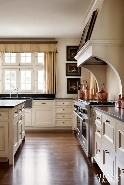 Cream kitchen with stainless steel range, dark countertops, and a large window with striped valance and gold curtains.