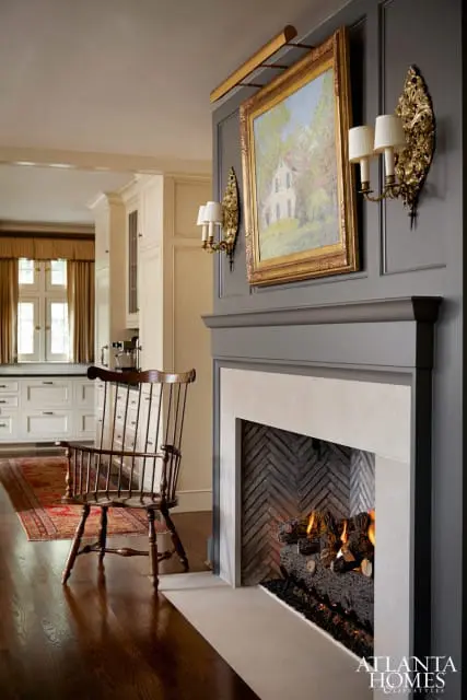 Living room with a gray fireplace, stone surround, herringbone brick firebox, and a framed painting above. A wooden Windsor chair sits on a rug in front of cream-colored cabinetry.