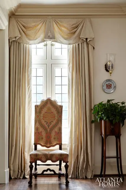 Traditional living room with a patterned upholstered armchair in front of a window with striped curtains. A wall sconce and plant stand are visible.