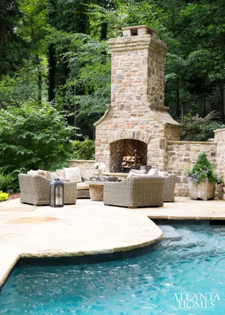 Stone outdoor fireplace and seating area next to a swimming pool, surrounded by lush green trees.