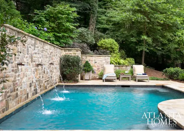 Inground swimming pool with a stone wall featuring multiple water spouts, surrounded by lush greenery and two lounge chairs.