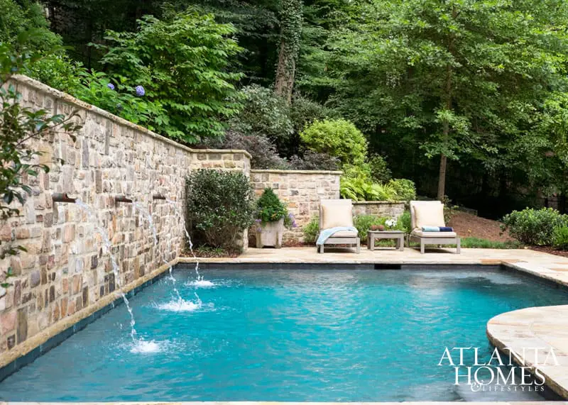 Inground swimming pool with a stone wall featuring multiple water spouts, surrounded by lush greenery and two lounge chairs.