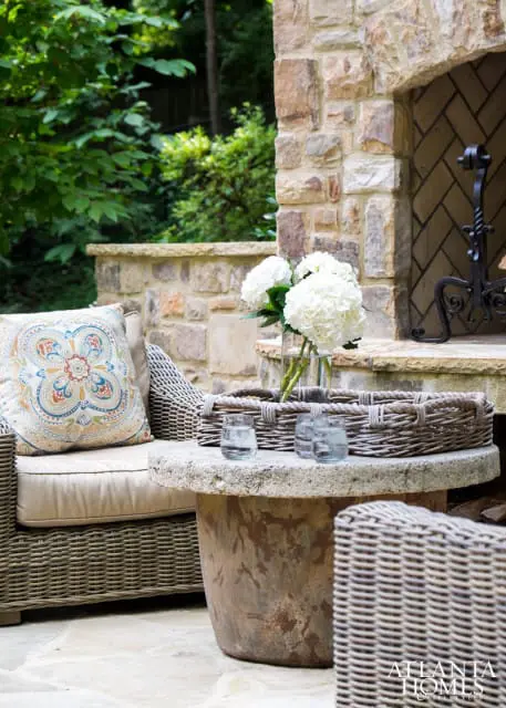 Wicker armchair with patterned cushion next to a stone table with hydrangea bouquet and woven trays. Stone fireplace in background.