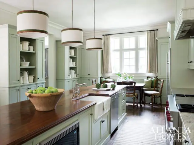Light green kitchen with a large wooden island topped with a bowl of green apples, white pendant lights, and a breakfast nook with a window.