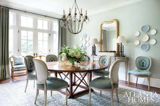 Light blue dining chairs with ornate green embroidery surround a polished wood dining table. A large gold mirror hangs above a console table with lamps.