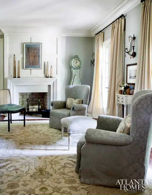 Living room with a fireplace, two upholstered armchairs, an ottoman, and a grandfather clock. A patterned rug covers the floor.