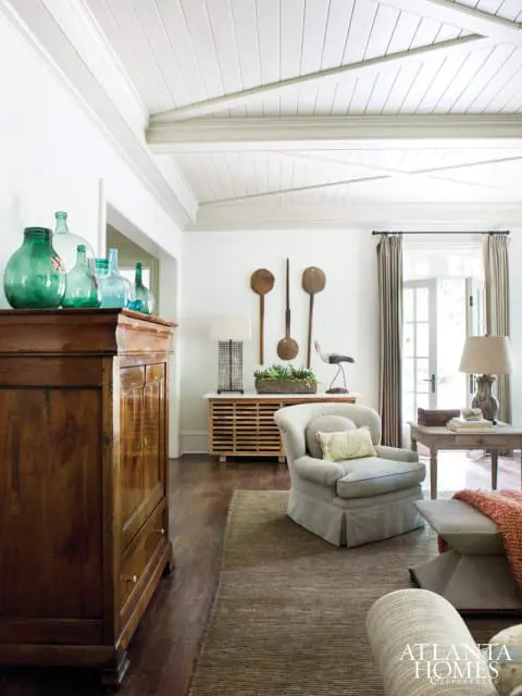 Living room with vaulted wood-paneled ceiling, antique wooden cabinet with green glass bottles, upholstered armchair, and a textured rug.