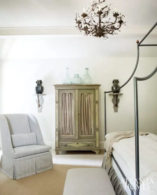 Bedroom with a light blue striped armchair, a distressed green armoire with draped fabric doors, and a metal canopy bed. A crystal chandelier hangs from the ceiling.
