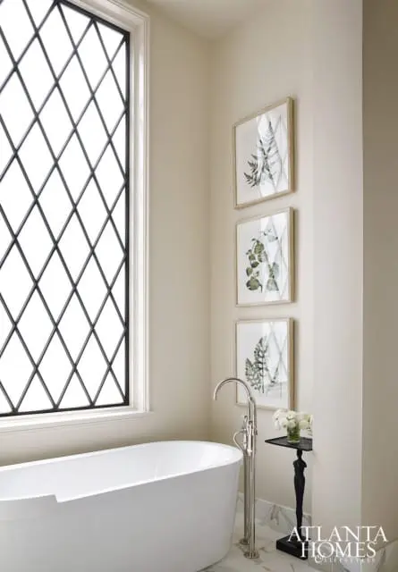 Cream bathroom with a white freestanding tub, diamond-paned window, three framed botanical prints, and a chrome floor-mounted faucet.