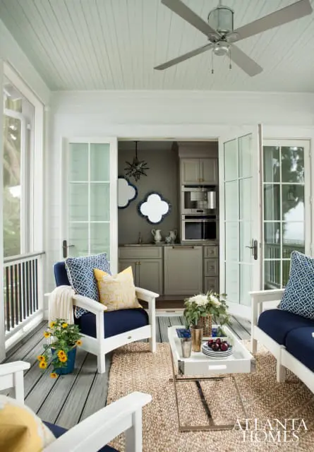 Covered porch with navy blue upholstered chairs, a white coffee table with a floral arrangement, and a small kitchen area visible through double doors.