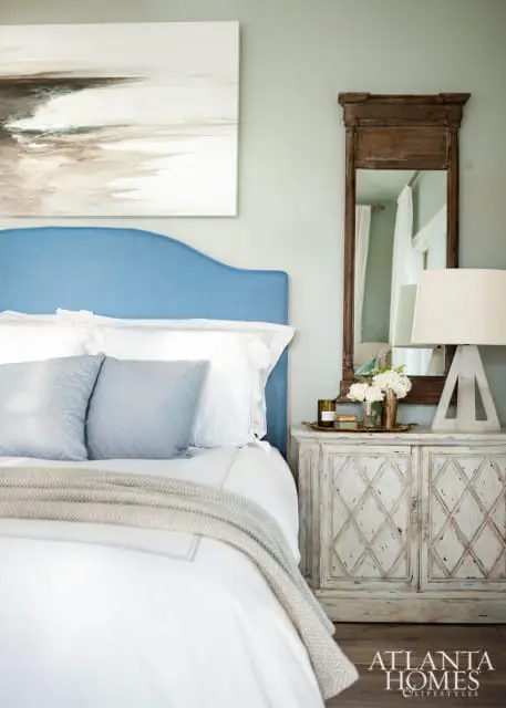 Bedroom with a blue upholstered headboard, white bedding, light blue accent pillows, and a distressed white nightstand with a wooden mirror above.