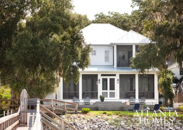 White coastal home with metal roof, screened porches, and a wooden boardwalk leading to the water.