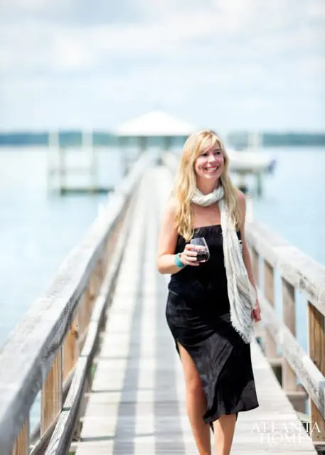 Woman in black dress and scarf walking on a wooden dock, holding a glass of red wine.