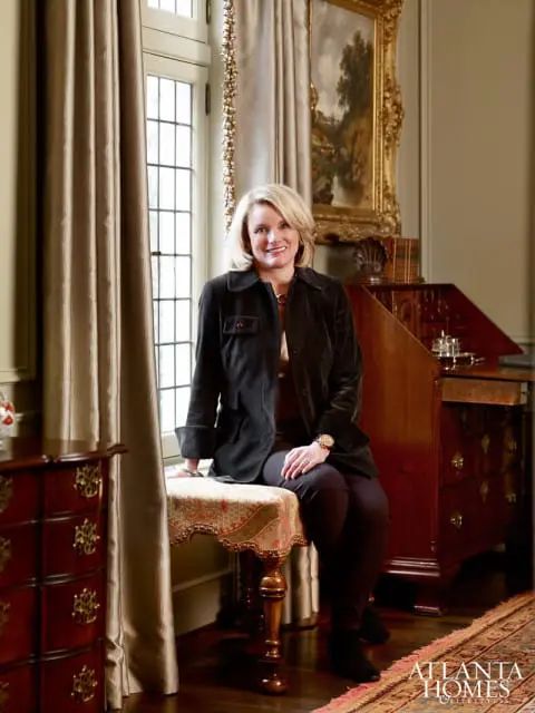 Woman in a dark jacket sitting on an upholstered bench in a room with a large window, antique desk, and ornate painting.
