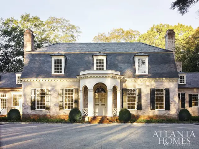 French-style brick home with a mansard roof, white-trimmed windows, black shutters, and a covered portico over the front door.