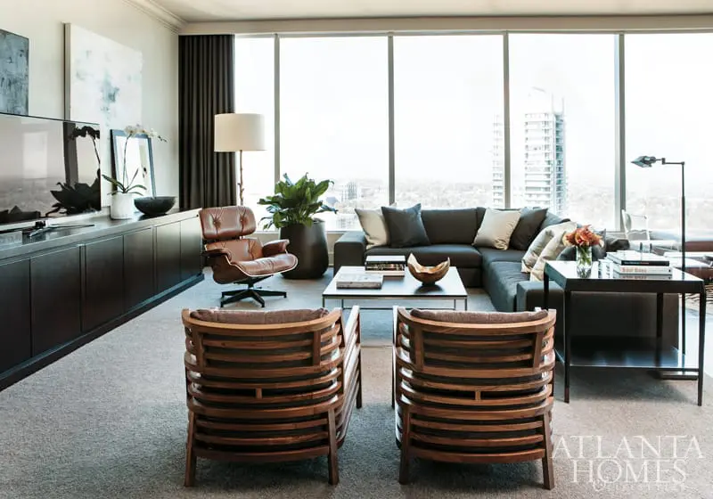 Modern living room with dark wood cabinetry, a brown leather lounge chair, a sectional sofa, and two wooden chairs facing a large window with city views.