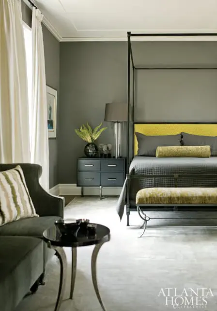 Bedroom with charcoal gray walls, a black canopy bed with a yellow geometric headboard, and dark gray nightstand with a glass lamp and vase of greenery.