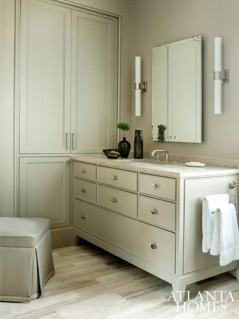 Bathroom with light gray cabinetry, marble countertop, and tiled floor. Features a tall cabinet, vanity with drawers, mirror, wall sconces, and a stool.