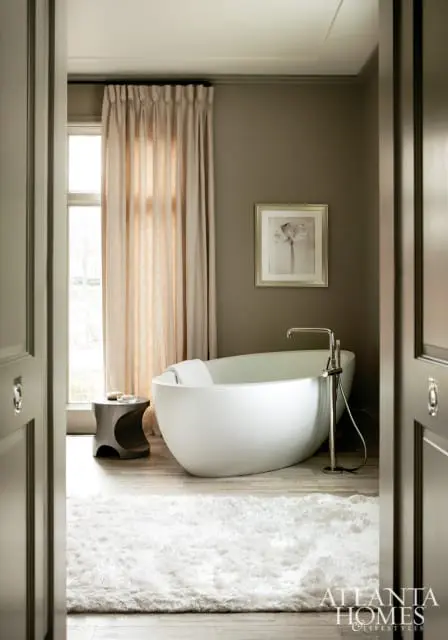 Modern bathroom with a freestanding white oval bathtub, chrome faucet, and a plush white rug on a light wood floor. A framed floral artwork hangs on the taupe wall.