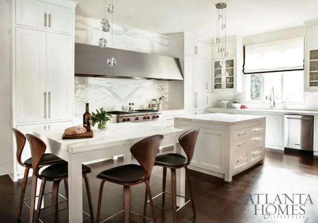 White kitchen with marble countertops and backsplash, stainless steel range hood, and a large island with seating.